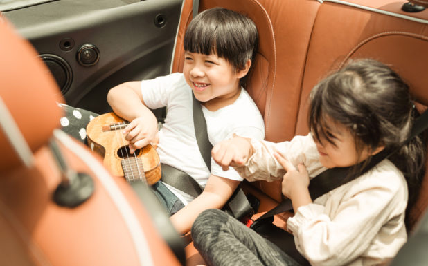 Two little kids smiling in the backseat of a vehicle; one with a ukulele.
