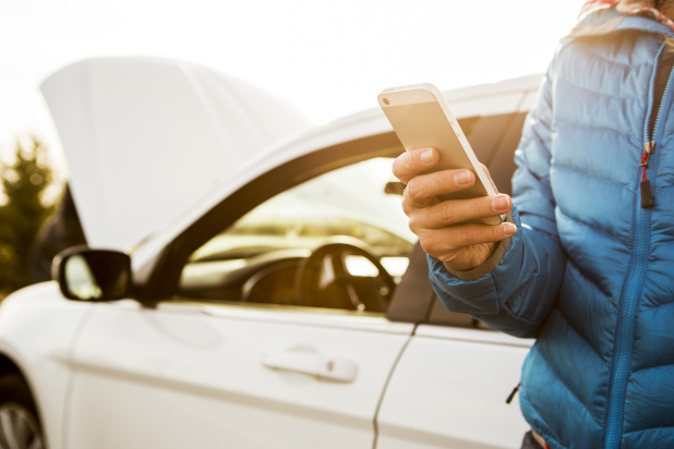 Photo of a person holding a mobile phone, beside a vehicle with its hood up.